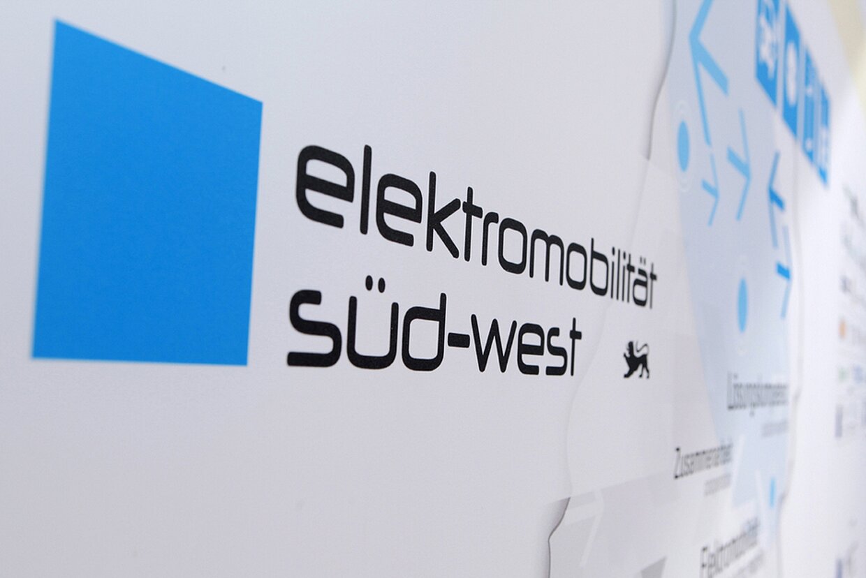 Wir freuen uns auf Ihren Besuch. Logo des Clusters Elektromobilität Süd-West von der Seite auf einer Wand fotografiert